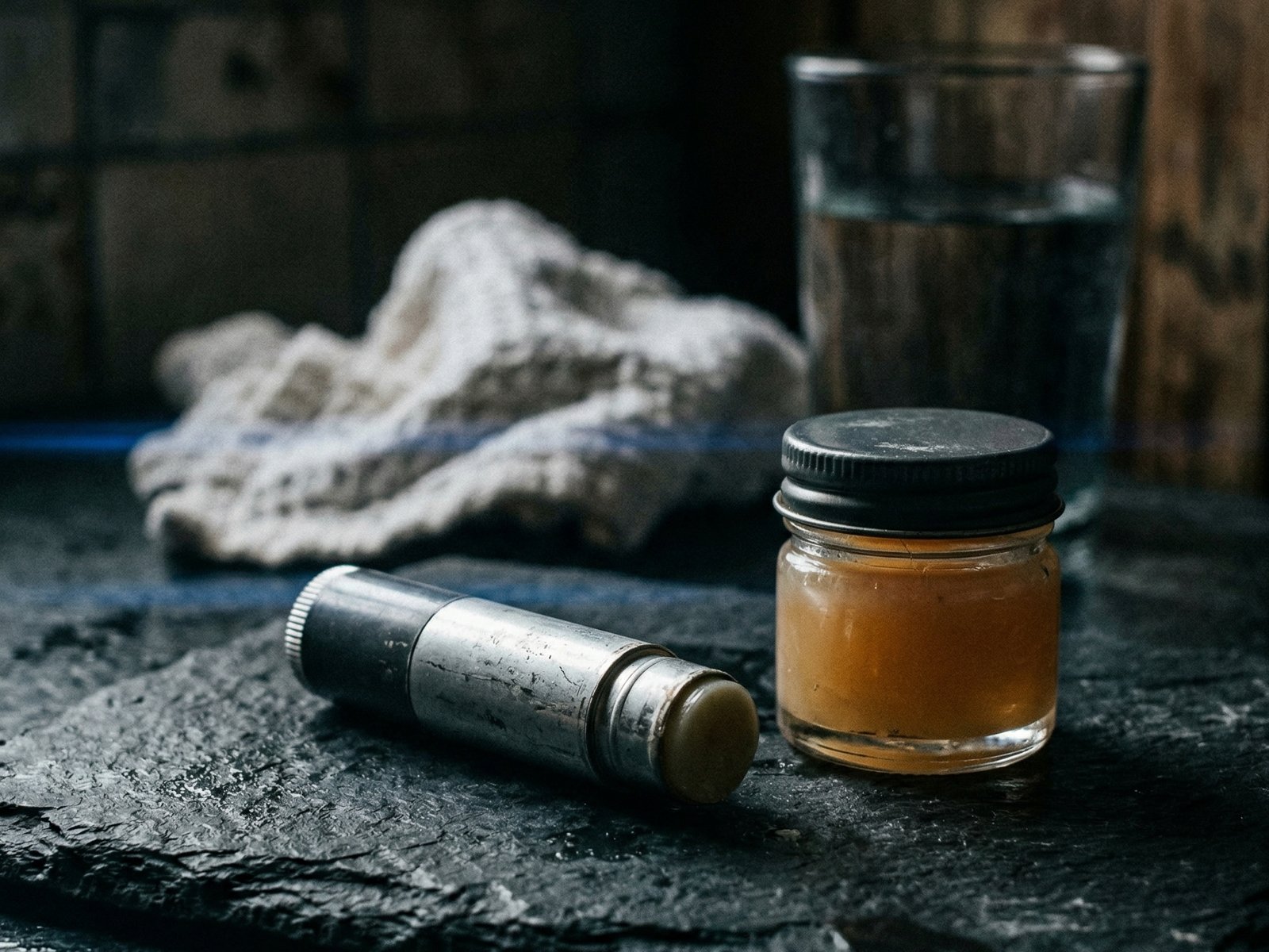 Lippenbalsam-Tube und Vaseline auf dunkler Schieferoberfläche, weisser Waschlappen und Wasserglas im Hintergrund